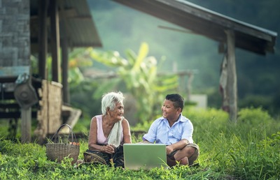 Idosa junto de seu cesto de vime e menino em frente de seu notebook conversam sorridentes, sentados lado a lado na vegetação rasa