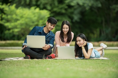 Um jovem e duas moças acomodados juntos no gramado sorriem diante de uma cena num dos notebooks Um jovem e duas moças acomodados juntos no gramado sorriem diante de uma cena num dos notebooks